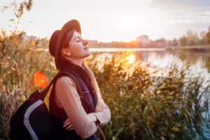 Traveler with backpack relaxing by autumn river at sunset. Young woman breathing deep feeling happy and free