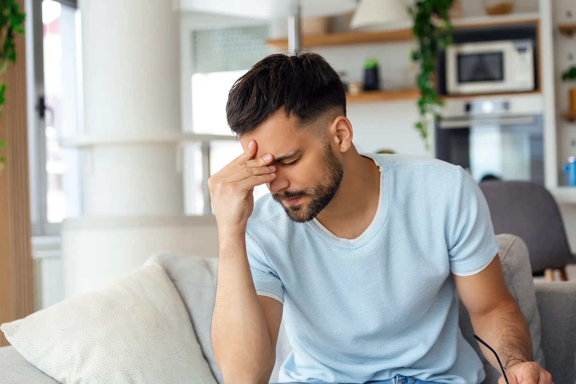 Fathers in recovery facing stress, sitting on a couch with hand on forehead in a thoughtful and emotional moment.