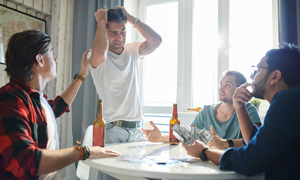 Friends playing cards, one man in white shirt looks frustrated while others smile, beer bottles on table.