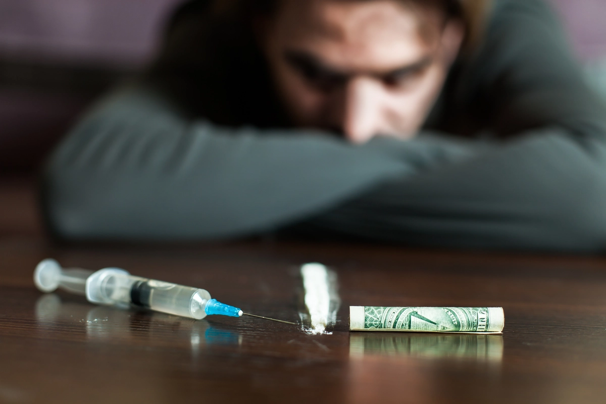 Person in gray shirt gazing at a table with a syringe, white powder, and rolled dollar bill.