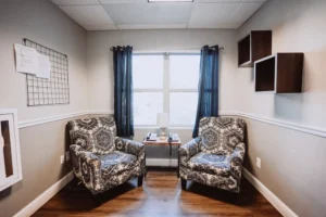 DSC09408-1-Recovered Two patterned armchairs in a cozy room with blue curtains and wall shelves.