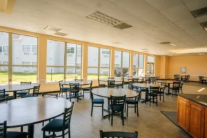 DSC09550-14 Spacious room with round tables, black chairs, large windows with outdoor view, and bright beige walls.