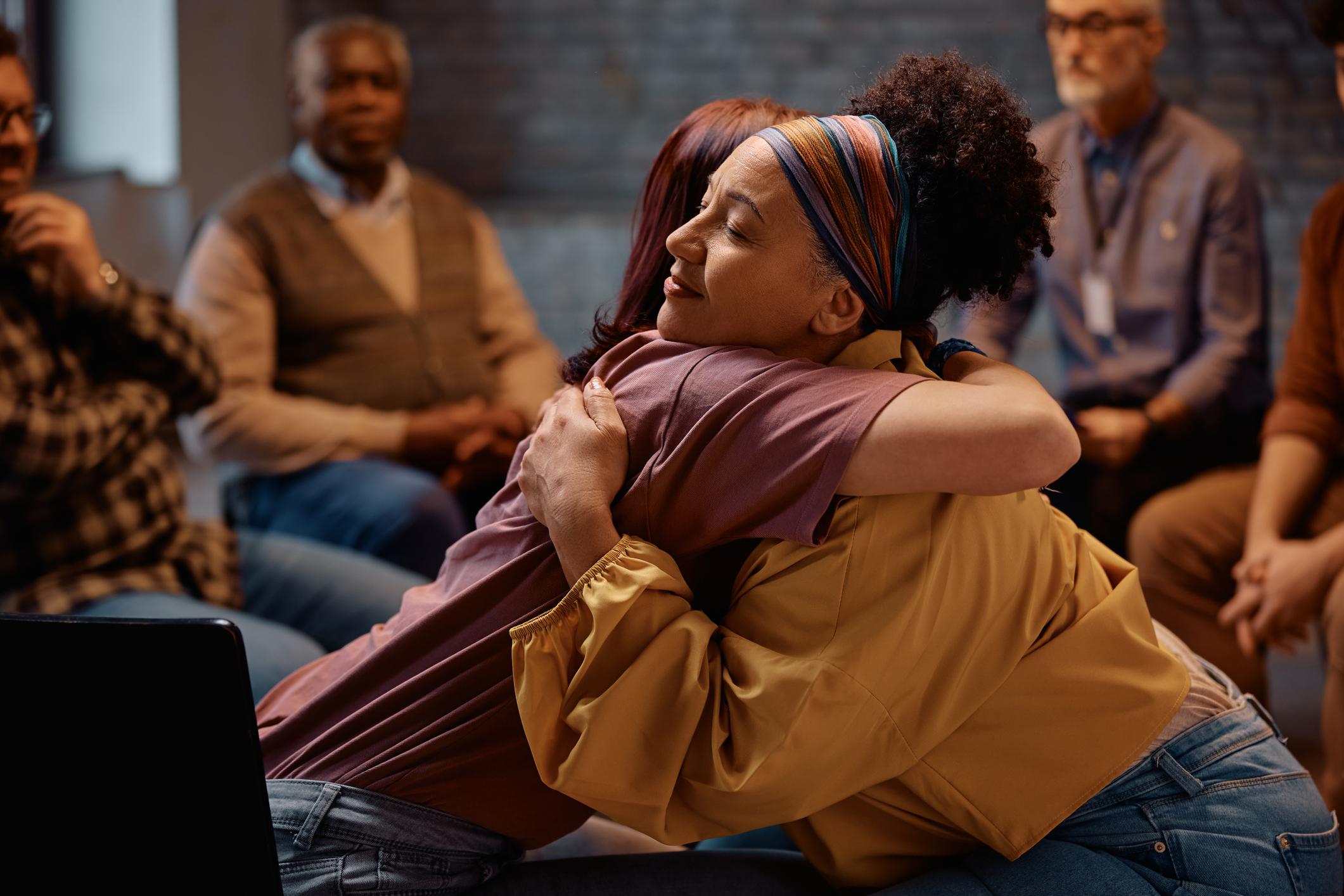 Supportive women embracing during group therapy at mental health center. Person wearing yellow shirt hugging another in a group setting.